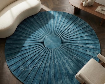 May include: A round, teal-colored rug with a radial pleated design. The rug's center is a solid teal circle, with radiating pleats extending outwards. The rug is placed on a dark wood floor, with a curved white sofa and wooden table in the background.
