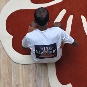 May include: Overhead view of a red and cream-colored rug with an organic shape. The rug is on a light-colored wooden floor. A person wearing a white t-shirt with the text "Rugs Mirzapur" is sitting on the rug.
