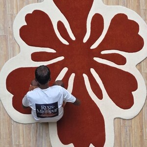 May include: A large, flower-shaped rug with a rust-colored center and off-white petals. The rug is on a light-colored wooden floor. A person wearing a white t-shirt with the text "Rugs Mirzapur" is sitting on the rug.
