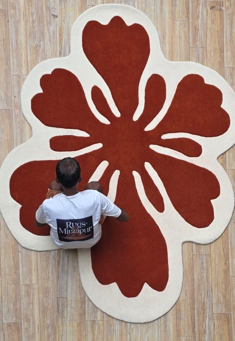 May include: A large, flower-shaped rug with a rust-colored center and off-white petals. The rug is on a light-colored wooden floor. A person wearing a white t-shirt with the text "Rugs Mirzapur" is sitting on the rug.