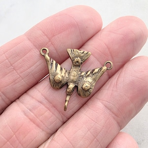 May include: A brass-toned metal bird charm featuring a stylized avian design with textured wings, a pointed tail, and a small circular loop on each side for attachment. The surface has a weathered, antique finish with visible dark patina.