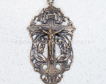 May include: A bronze-colored metal crucifix with a detailed design. The crucifix features a figure of Jesus Christ on the cross, with two figures kneeling in prayer on either side. The crucifix has a loop at the top for hanging.