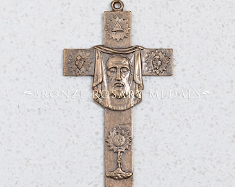 May include: A bronze religious cross with a detailed image of Jesus' face, surrounded by a crown of thorns and a cloth. The cross also features a triangle with a circle inside, a heart with a cross inside, and a sun with a cross inside.