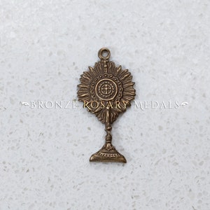 May include: A bronze metal religious charm featuring a monstrance with a cross and a sunburst design. The charm is on a white background.