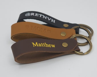 May include: Three leather keychains with metal key rings. The keychains are brown, tan, and black. The brown keychain has the name "Matthew" embossed in gold lettering. The tan keychain has the word "MOM" embossed in gold lettering. The black keychain has the text "@RETNVH" embossed in silver lettering.