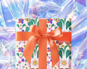 May include: A square gift box wrapped in white paper featuring a colorful botanical and celestial pattern of green leaves, pink flowers, mushrooms, and suns with eyes. The box is tied with a wide, solid orange satin ribbon tied in a bow.