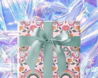May include: A square gift box wrapped in light pink paper featuring a repeating pattern of butterflies, flowers, rainbows, and eyes, tied with a wide sage green satin ribbon that is knotted into a bow centered on the front of the package.