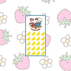 May include: A rectangular card with a blue border featuring the text "Tis the Seasoning" and illustrations of a lobster, pot, and lemons. The card is set against a white background with pink strawberries and white flowers.