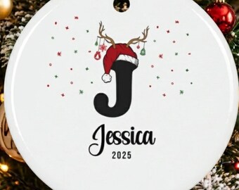 May include: A round white ceramic Christmas ornament featuring a large black letter J topped with a red Santa hat and reindeer antlers. The name Jessica is printed in black script below the letter, followed by the year 2025 in black text.