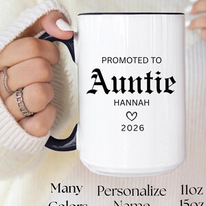 May include: White ceramic mug with a black handle and rim. The mug features the text "PROMOTED TO Auntie HANNAH 2026" in black. Additional text includes "Many Colors", "Personalize Name", "11oz", "15oz", and "Limited Edition". The mug has a heart symbol.