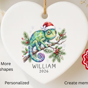 May include: White heart-shaped Christmas ornament with a chameleon wearing a Santa hat, on a branch with berries and snowflakes. The ornament has the name "WILLIAM" and the year "2026" printed on it.