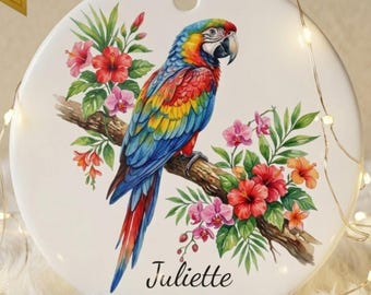 May include: A round white ceramic ornament featuring a colorful macaw parrot perched on a branch surrounded by red hibiscus and pink orchid flowers. The name Juliette and the year 2026 are printed in black script below the bird on the ornament.