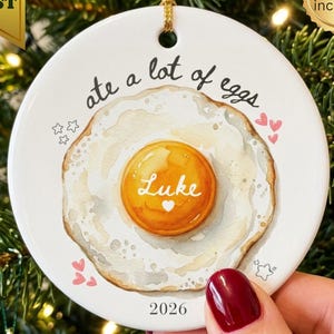 Può includere: Un ornamento rotondo in ceramica bianca con un'illustrazione ad acquerello di un uovo al tegamino e il testo "ate a lot of eggs" sopra. Il tuorlo dell'uovo riporta il nome "Luke" e un piccolo cuore. L'anno "2026" è stampato in basso.