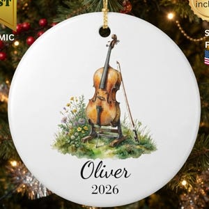 May include: A round white ceramic ornament featuring a watercolour illustration of a brown cello on a stand with a bow and colourful wildflowers. The name Oliver and the year 2026 are printed in black script and serif font below the illustration.