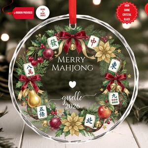 May include: A clear, round Christmas ornament with a festive wreath design. The wreath includes red bows, gold bells, and Mahjong tiles. The text "MERRY MAHJONG" is displayed, along with a name and the year 2026.
