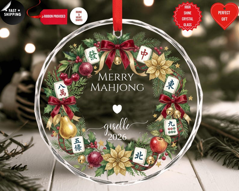 May include: A clear, round Christmas ornament with a festive wreath design. The wreath includes red bows, gold bells, and Mahjong tiles. The text "MERRY MAHJONG" is displayed, along with a name and the year 2026.