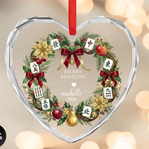 May include: Heart-shaped glass ornament with a festive Mahjong-themed design. The ornament features a wreath of greenery, red bows, and Mahjong tiles. The text "MERRY MAHJONG" and a name and year are printed in the center.
