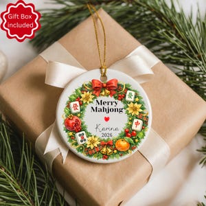 Pu&ograve; includere: Ornamento natalizio in ceramica bianca con un filo dorato, con un design a ghirlanda con la scritta "Merry Mahjong" e il nome "Karina" con l'anno 2026. La ghirlanda include fiocchi rossi, fiori e tessere di Mahjong. L'ornamento &egrave; su un regalo confezionato.