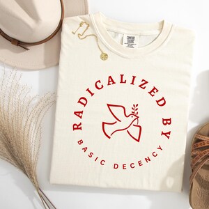 以下が含まれることがあります： 「RADICALIZED BY BASIC DECENCY」の赤い文字が、オリーブの枝を持った鳩を囲むクリーム色のTシャツ。茶色のサンダル、ベージュの帽子、金のネックレスも写っています。