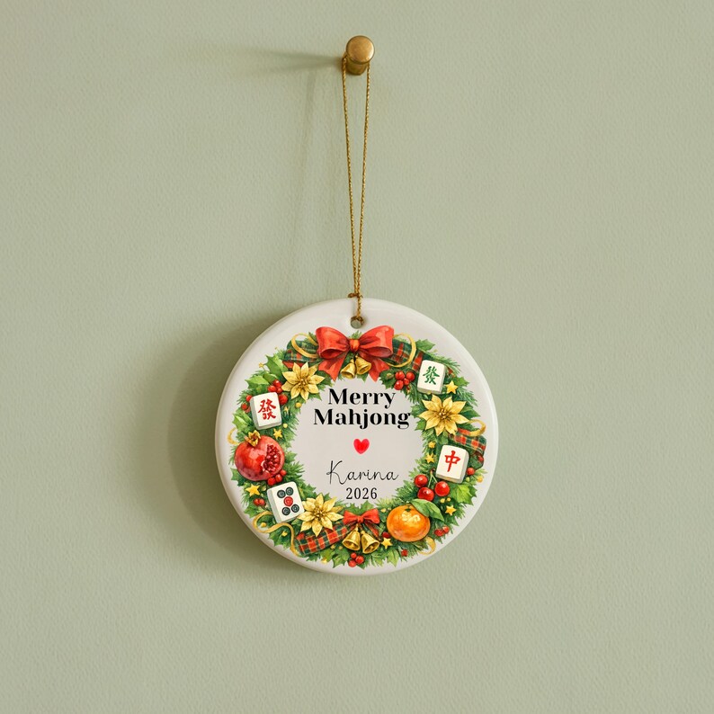 Pu&ograve; includere: Ornamento natalizio rotondo in ceramica bianca con un design a ghirlanda festiva. La ghirlanda presenta fiocchi rossi, fiori gialli e tessere di Mahjong. Il testo "Merry Mahjong" e un nome sono visualizzati. Un filo d'oro &egrave; attaccato per appenderlo.