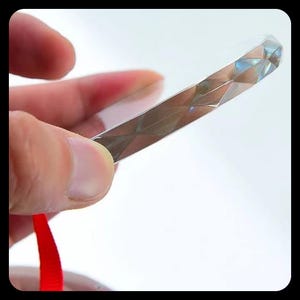 May include: A clear, rectangular prism with a faceted, diamond-like design. The prism is held by a hand, with a red ribbon visible at the bottom. The object reflects light, creating a shimmering effect.