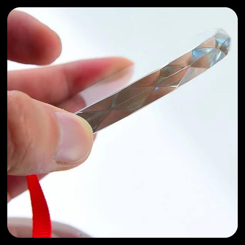 May include: A clear, rectangular prism with a faceted, diamond-like design. The prism is held by a hand, with a red ribbon visible at the bottom. The object reflects light, creating a shimmering effect.