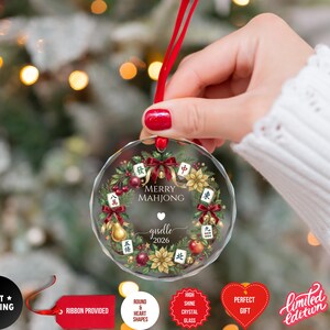 May include: A clear, round Christmas ornament with a red ribbon. The ornament features a wreath design with Mahjong tiles, berries, and the text "MERRY MAHJONG" and "giselle 2026".