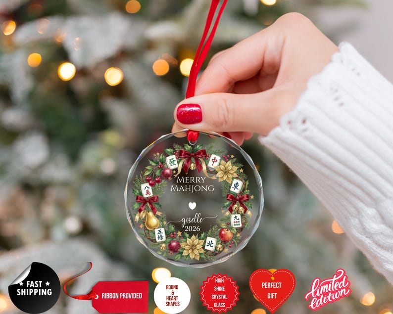 May include: A clear, round Christmas ornament with a red ribbon. The ornament features a wreath design with Mahjong tiles, berries, and the text "MERRY MAHJONG" and "giselle 2026".