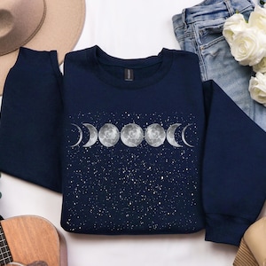 Puede incluir: Sudadera azul marino con un diseño de fases lunares blancas sobre un fondo azul oscuro con motas blancas. El diseño presenta una luna creciente, una media luna, una luna llena y una luna gibosa menguante.