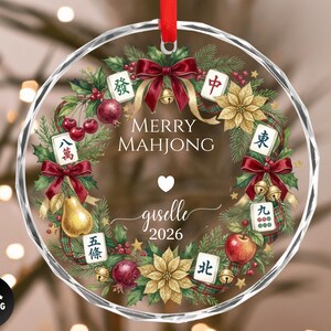 May include: A clear, round Christmas ornament with a red ribbon. The ornament features a wreath design with Mahjong tiles, fruit, and the text "MERRY MAHJONG" and "giselle 2026".