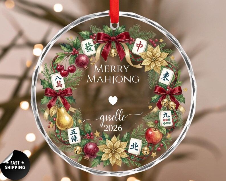 May include: A clear, round Christmas ornament with a red ribbon. The ornament features a wreath design with Mahjong tiles, fruit, and the text "MERRY MAHJONG" and "giselle 2026".