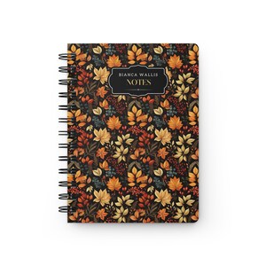 May include: A black spiral notebook with a fall-themed design. The cover features a pattern of orange, yellow, and red leaves, berries, and branches on a black background. The text "Bianca Wallis NOTES" is printed in gold on a black label.