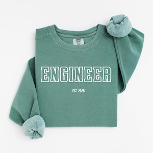 Puede incluir: Una sudadera verde con la palabra "ENGINEER" impresa en blanco en la parte delantera. El texto está en una fuente en negrita y en bloque. Debajo de la palabra "ENGINEER" está el texto "EST. 2025".