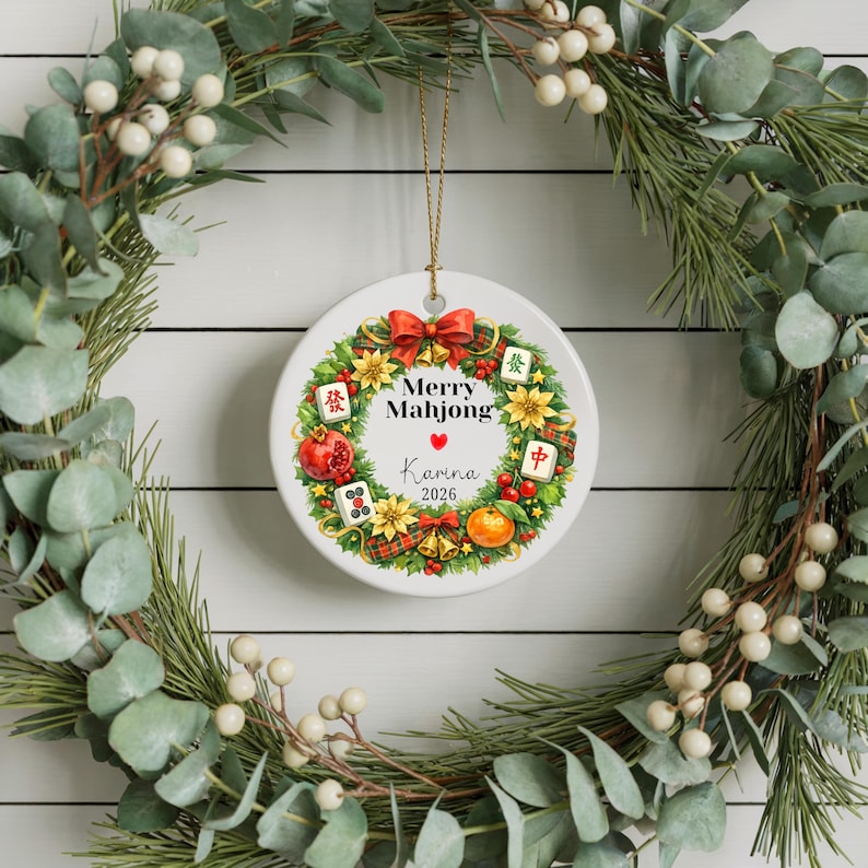 Pu&ograve; includere: Un ornamento natalizio rotondo e bianco con un filo dorato, con un design "Merry Mahjong". L'ornamento include una ghirlanda di fiori, frutta e tessere di Mahjong. Il nome "Karina" e l'anno "2026" sono stampati.