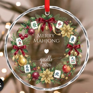 May include: A clear, round Christmas ornament with a decorative wreath design. The wreath includes red bows, gold bells, and Mahjong tiles. The text "Merry Mahjong" and a name and year are in the center. A red ribbon is at the top.