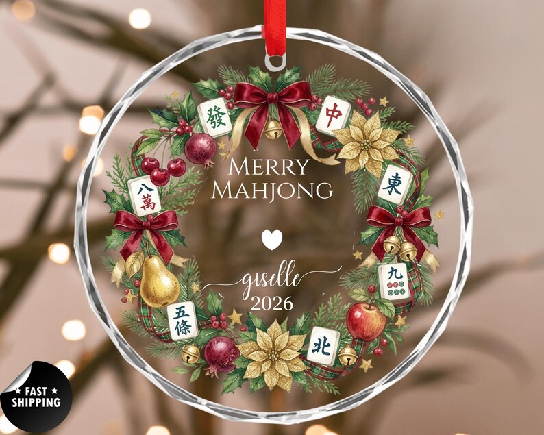 May include: A clear, round Christmas ornament with a decorative wreath design. The wreath includes red bows, gold bells, and Mahjong tiles. The text "Merry Mahjong" and a name and year are in the center. A red ribbon is at the top.