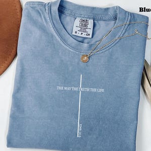 以下が含まれることがあります： 「THE WAY THE TRUTH THE LIFE」と「JOHN 14:6」の文字が白でプリントされた、ブルーデニムカラーのTシャツ。 金色のペンダント付きネックレスがシャツの上にかけられています。 シャツは折りたたまれており、「COMFORT COLORS」と書かれたタグが付いています。