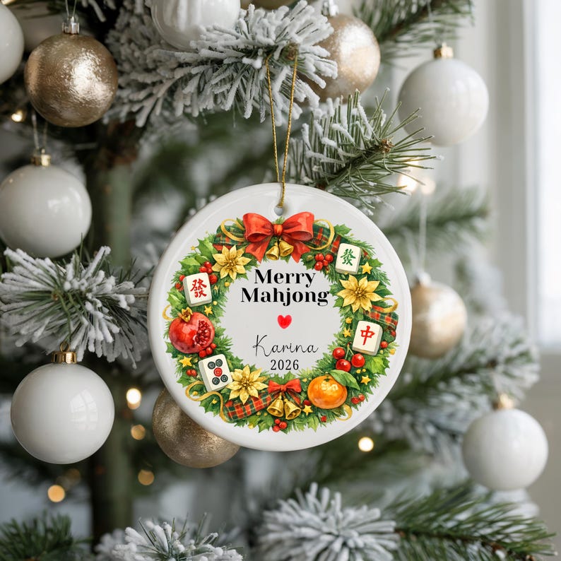 Pu&ograve; includere: Un ornamento natalizio in ceramica bianca con un filo d'oro, con un design a ghirlanda con le parole "Merry Mahjong" e "Karina 2026". La ghirlanda include un fiocco rosso, fiori e tessere di Mahjong. L'ornamento &egrave; appeso a un albero di Natale.