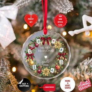 May include: A clear, round Christmas ornament with a red ribbon. The ornament features a wreath design with Mahjong tiles, red bows, and the text "Merry Mahjong" and "giselle 2026".