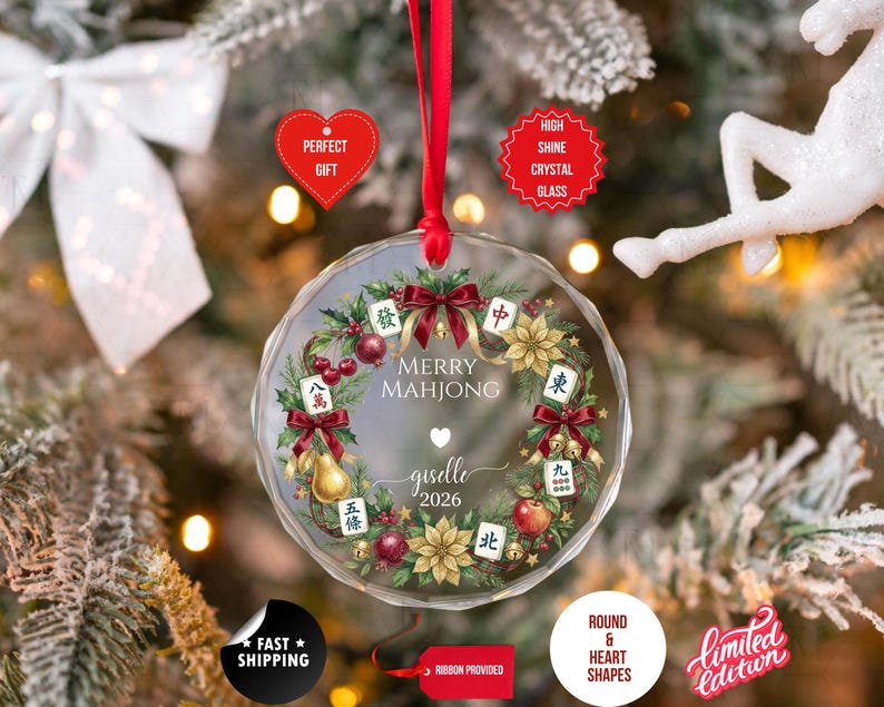 May include: A clear, round Christmas ornament with a red ribbon. The ornament features a wreath design with Mahjong tiles, red bows, and the text "Merry Mahjong" and "giselle 2026".