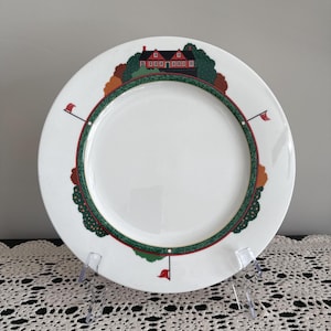 May include: White ceramic plate with a decorative border featuring a red-roofed house, trees, and golf flags. The border is green and red, with a white center. The plate is displayed on a clear plastic stand.