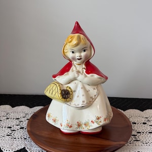 May include: A ceramic figurine of Little Red Riding Hood, featuring a red hooded cape, a white dress with floral accents, and a yellow basket. The figurine is standing on a wooden plate.