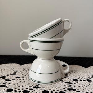 May include: Three white ceramic teacups stacked on top of each other. Each cup has a handle and is decorated with two thin, dark green horizontal stripes. The cups are arranged on a white lace doily.
