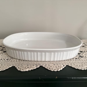 May include: White oval baking dish with a ribbed exterior. The dish is made of ceramic and sits on a decorative, cream-colored lace doily. The dish is empty and ready for use.
