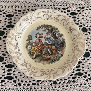 May include: A vintage decorative plate with a scalloped edge and gold leaf accents. The center features a pastoral scene with figures in period clothing. The plate is cream-colored with a diameter of approximately 7 inches.