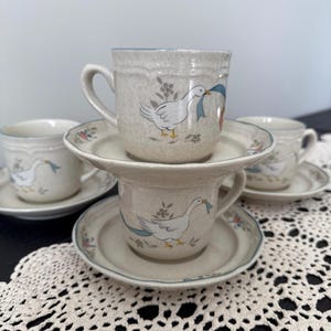 May include: A set of four beige ceramic mugs and saucers. Each mug features a white goose with a blue ribbon and floral accents. The saucers have a matching floral pattern around the edges. The mugs are stacked for display.
