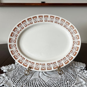 May include: A white oval serving platter with a brown geometric pattern around the edge. The platter is on a white crocheted doily.