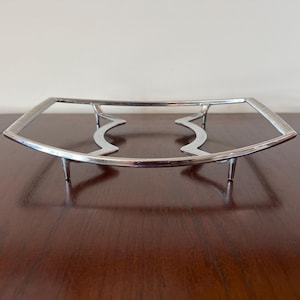 May include: A silver metal serving stand with a curved design and four legs. The stand is designed to hold a plate or bowl.