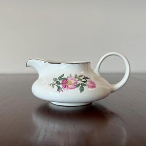 May include: A white ceramic creamer with a silver rim and a floral design featuring pink roses and green leaves.