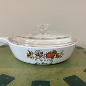 May include: A white casserole dish with a clear glass lid. The dish features a colorful illustration of vegetables, including a tomato and mushrooms. The text "Les Persil" is printed on the front.